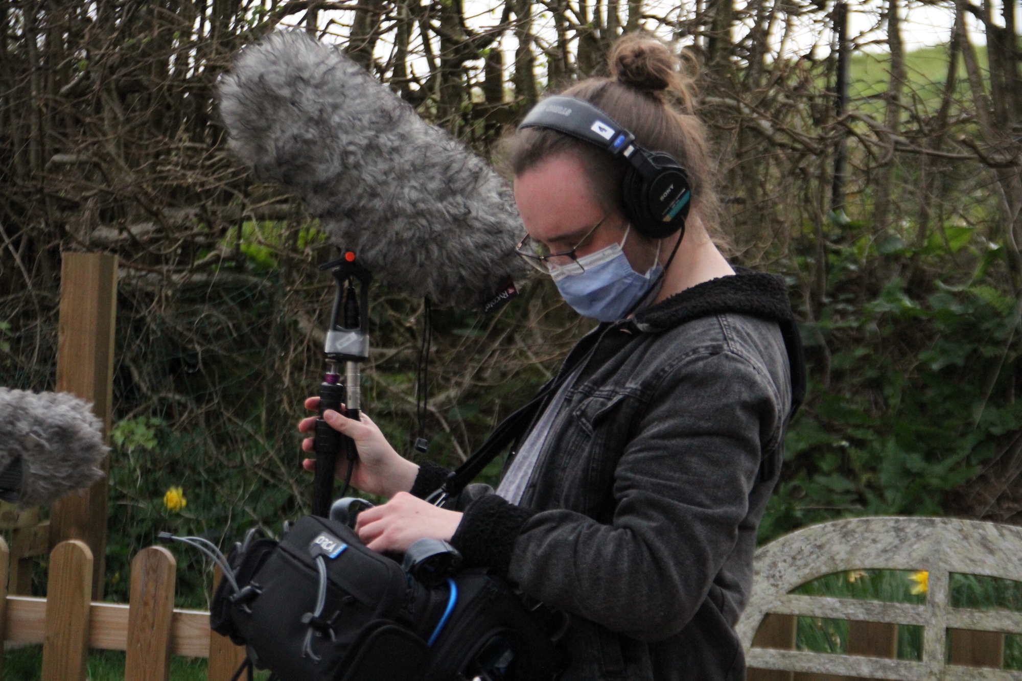 A person wearing earphones and a face mask stands looking down while holding a boom microphone