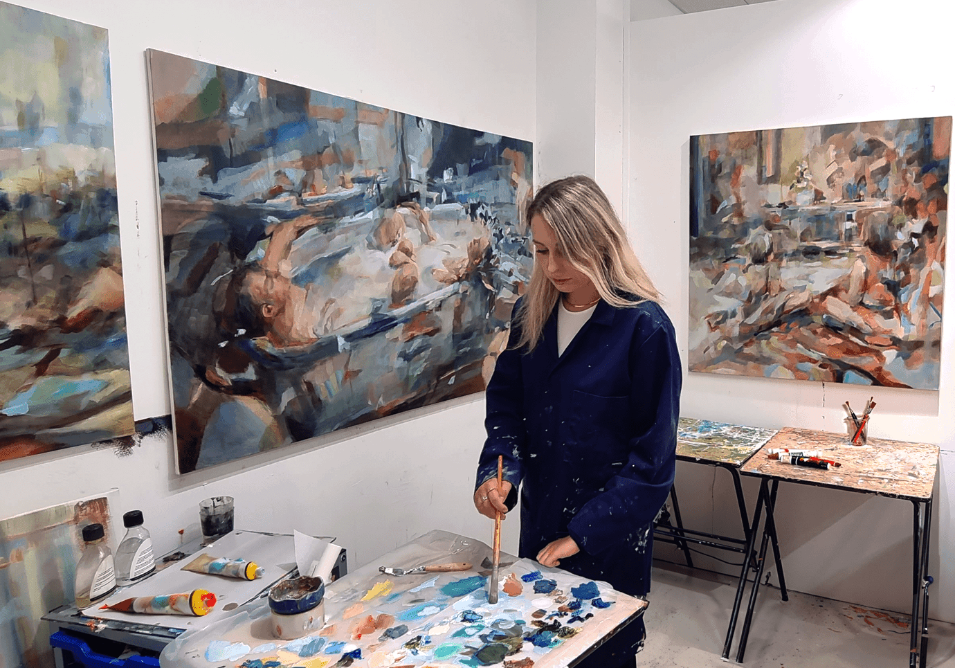 Painter E Shepard standing at a table working, with various paintings on the walls around them.