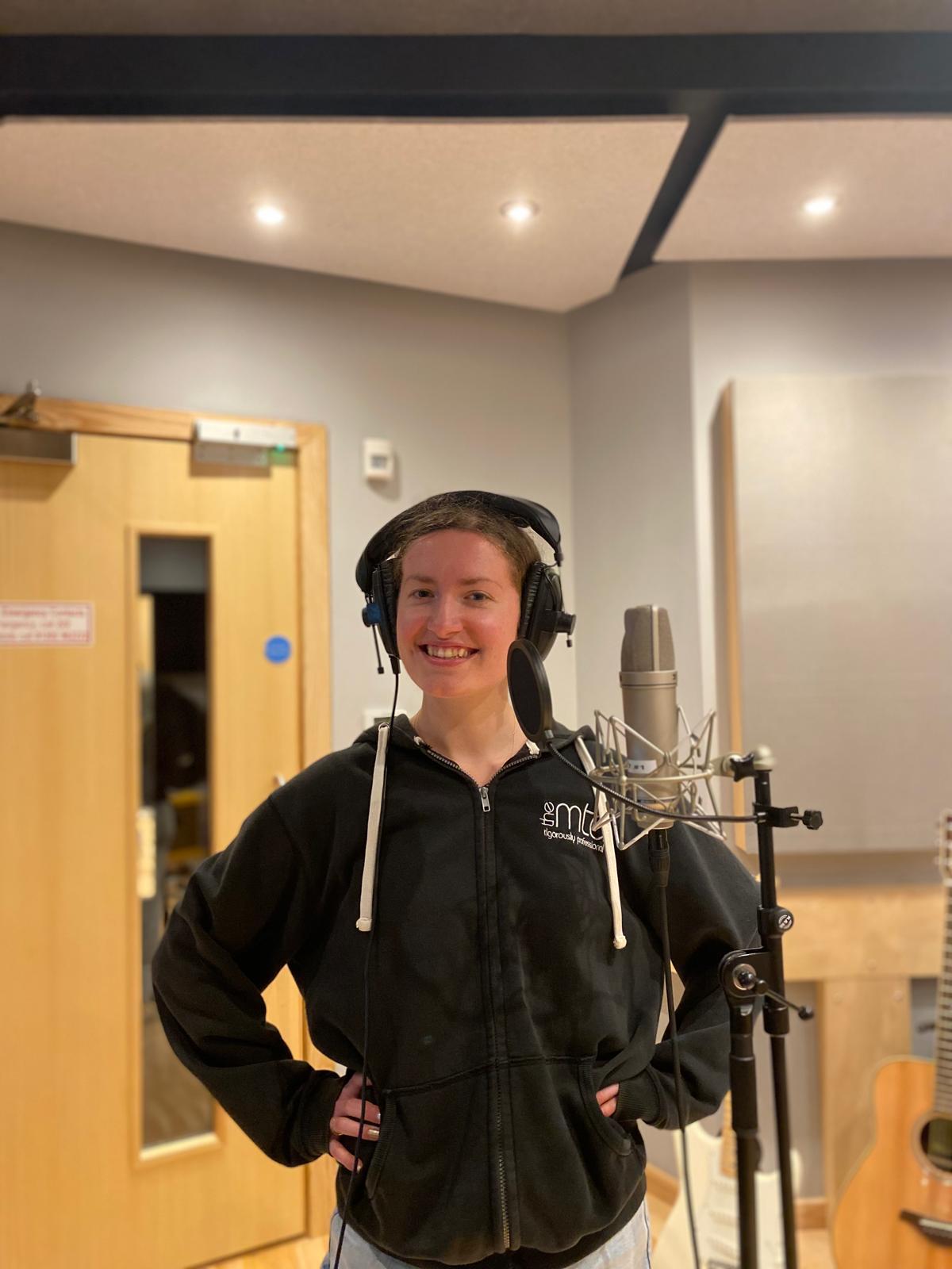 E. Shipley in a practice room stood by a mic wearing headphones.