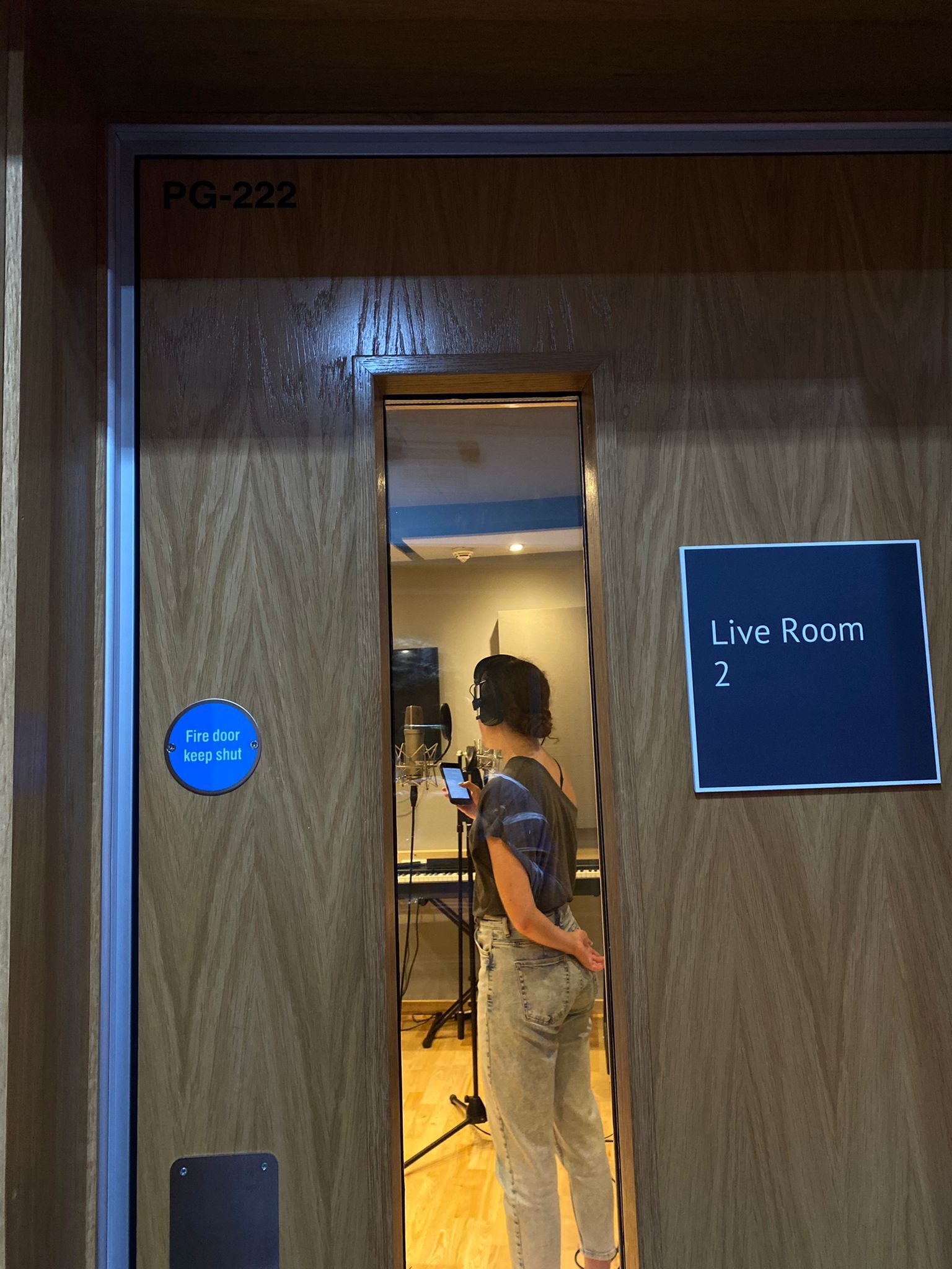 Ella Shipley through a practice room door stood by a mic wearing headphones.