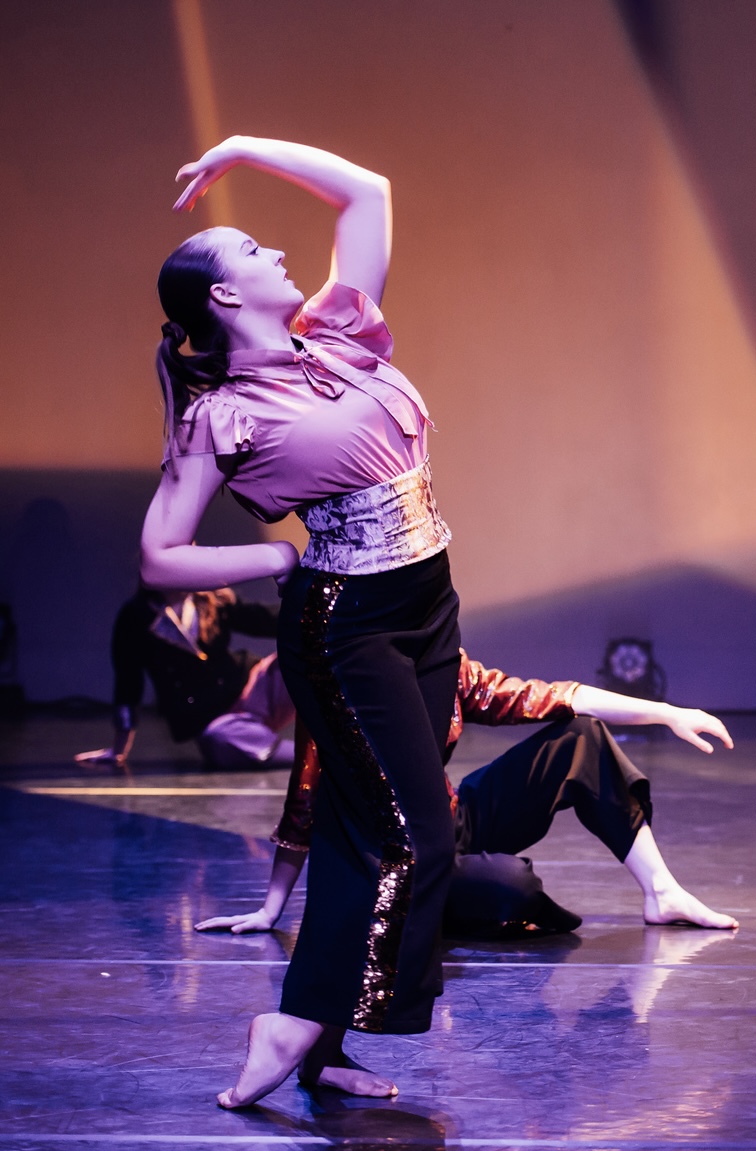 A dancer in a pink top and sequined black trousers performs on stage, with one arm raised elegantly. In the background, two other dancers are positioned on the floor, dressed in black and red outfits, against a softly lit backdrop.