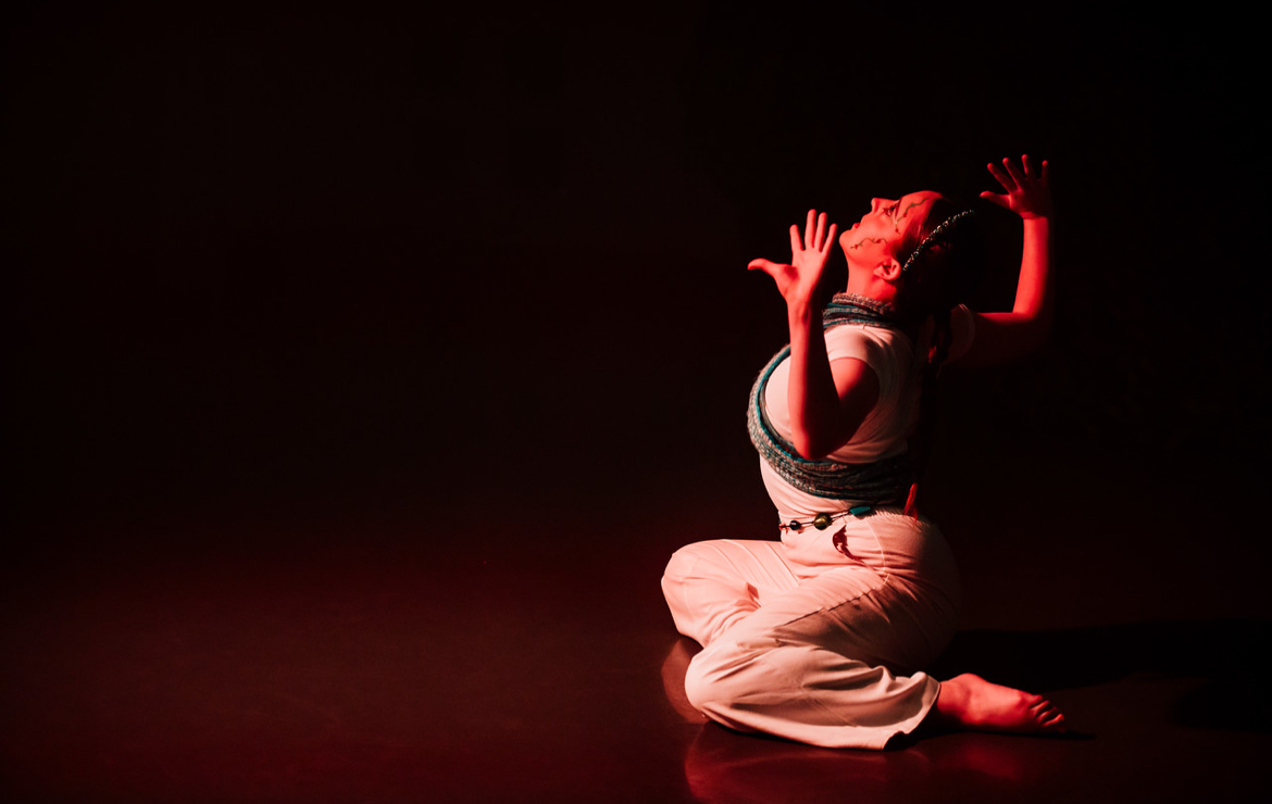 A person dressed in white clothing kneels on the floor with arms raised, surrounded by a dark background illuminated by red lighting. The focus is on the figure's dynamic pose and the contrasting light.