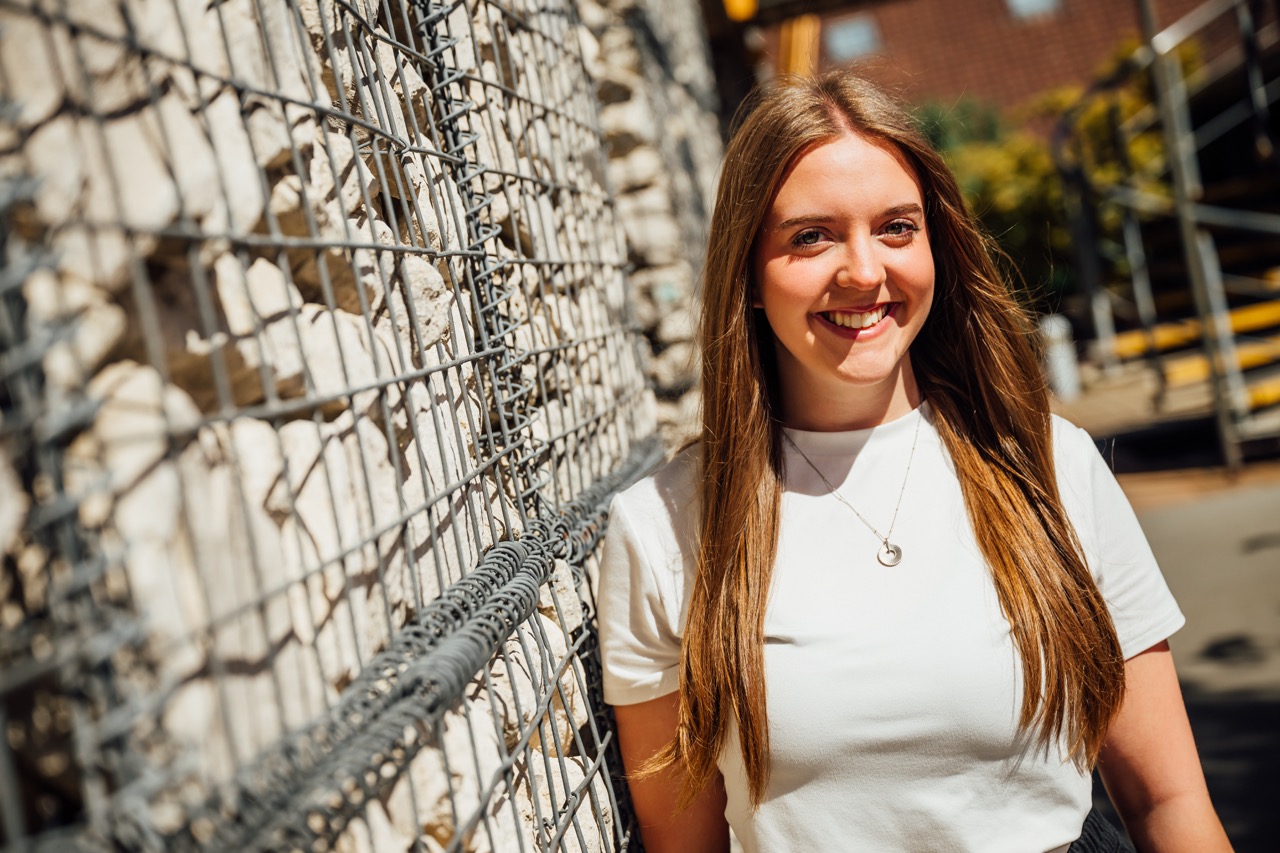 A young woman with long brown hair is standing against a stone wall, wearing a white t-shirt and a silver necklace. The sunlight illuminates the scene, highlighting her casual, outdoor setting.