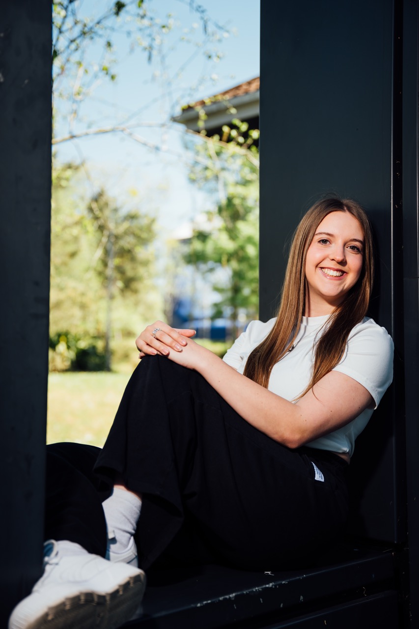 A young woman with long hair sits on a black structure, wearing a light blue top and black trousers. She rests her arms on her knees, with trees and a building visible in the background under a clear sky.