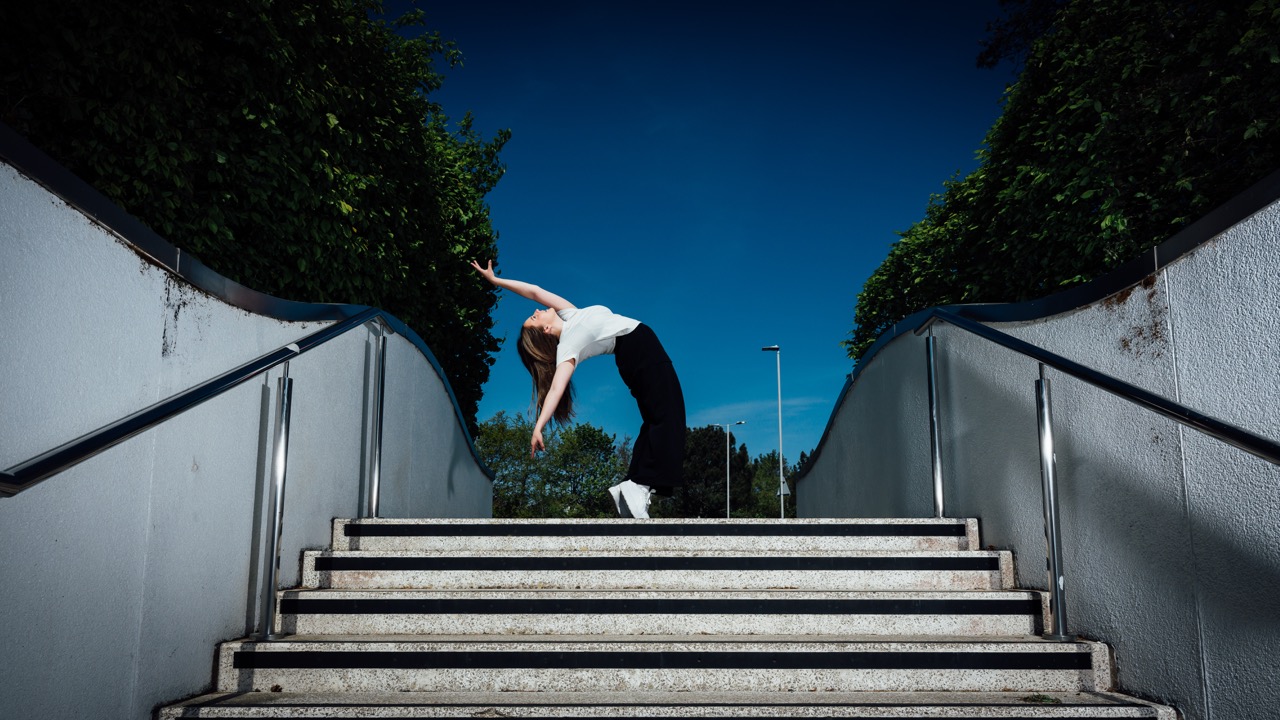 A person performing a dance move while standing on a staircase. The individual, wearing a white top and black trousers, leans back, creating a dynamic pose against a backdrop of clear blue sky and greenery.