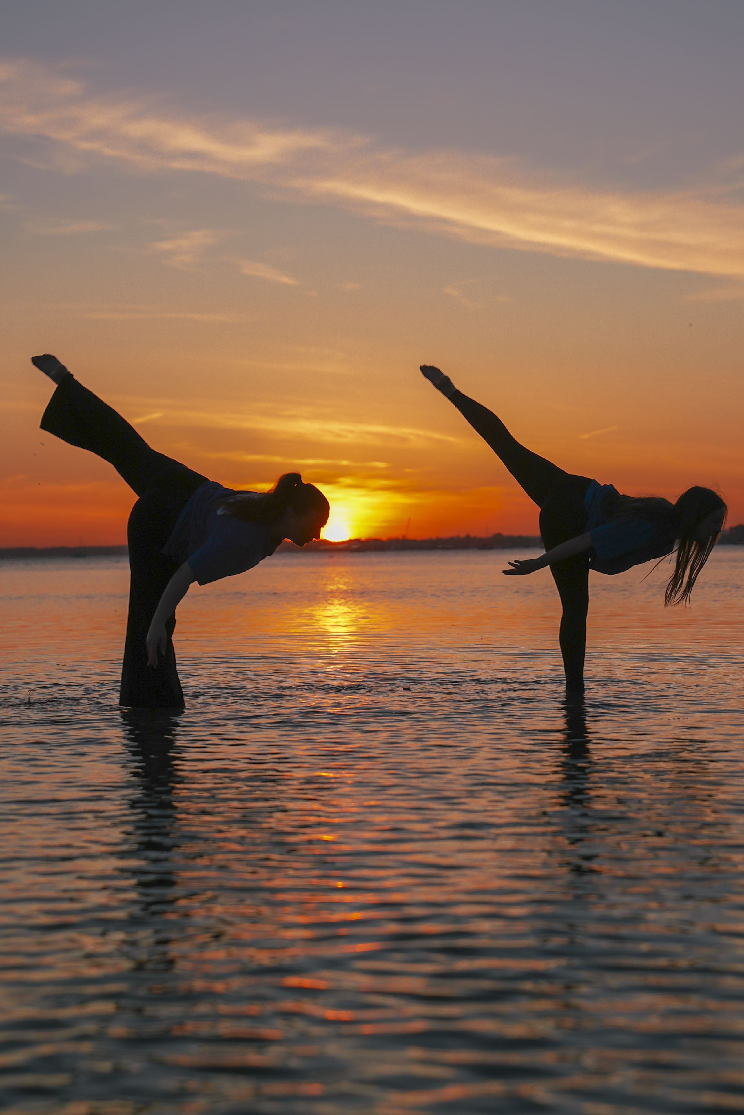 Two dancers perform on the surface of a body of water at sunset. One dancer bends forward with one leg raised, while the other stands on one leg, extending their arms, against a backdrop of orange and purple hues in the sky.