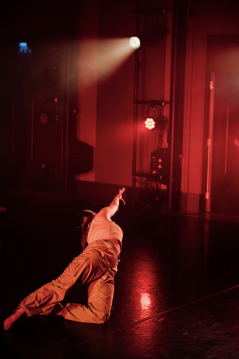 A dancer in beige trousers and a white shirt is lying on the floor, with one arm extended upwards. The scene is illuminated by red stage lights, creating a dramatic atmosphere, while the background features darkened curtains and stage equipment.