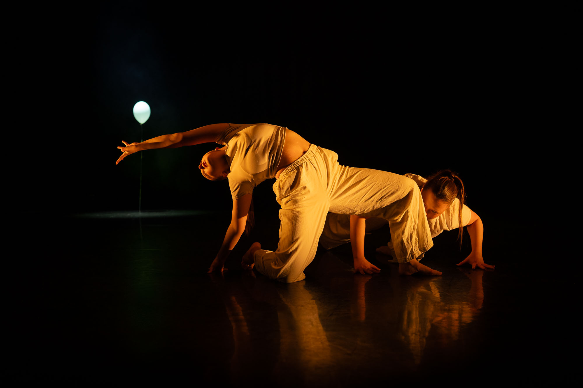 Two dancers in white outfits performing on a dimly lit stage. One dancer is arching backwards while the other moves on all fours beside her. A single illuminated balloon stands in the background, adding a soft light to the scene.
