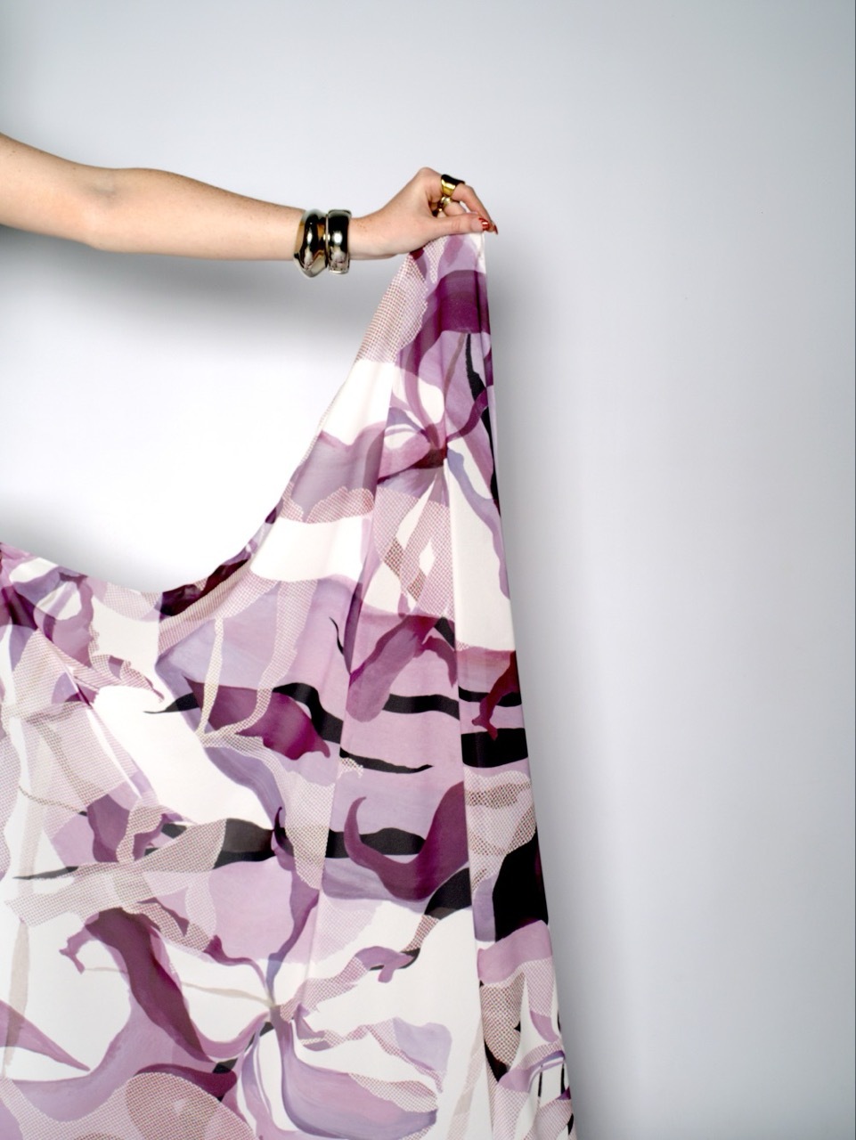 Hand wearing black bracelets holds pink and purple camouflage-patterned fabric against white wall.