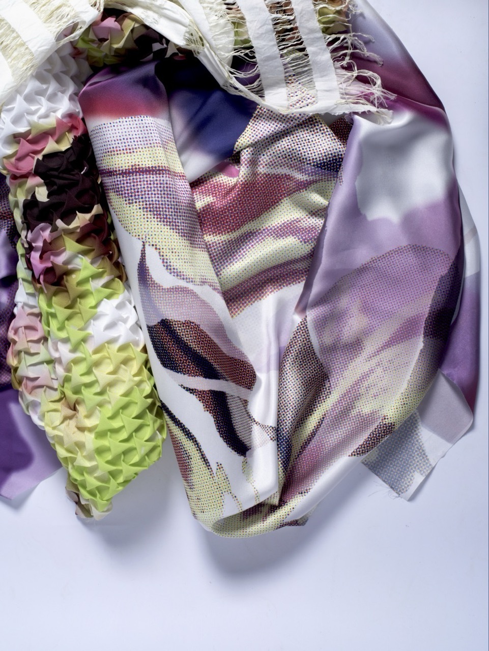 Crumpled mesh fabric in purple, white, and multicoloured patterns with textured green and patterned material pieces on white surface.