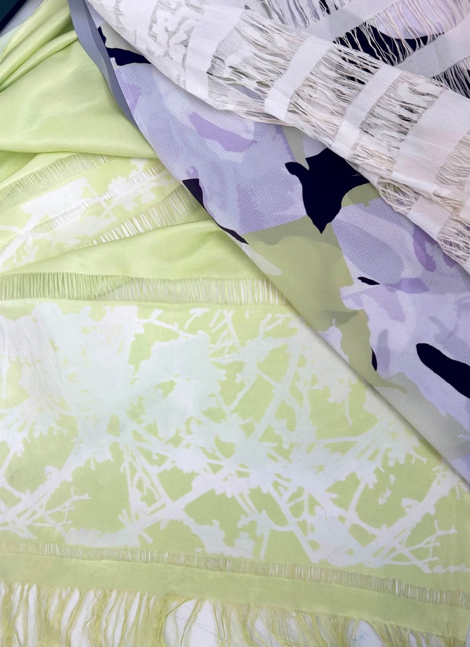 Layered fabrics in pale green, lavender, and white with fringe edges and black floral patterns overlapping diagonally.