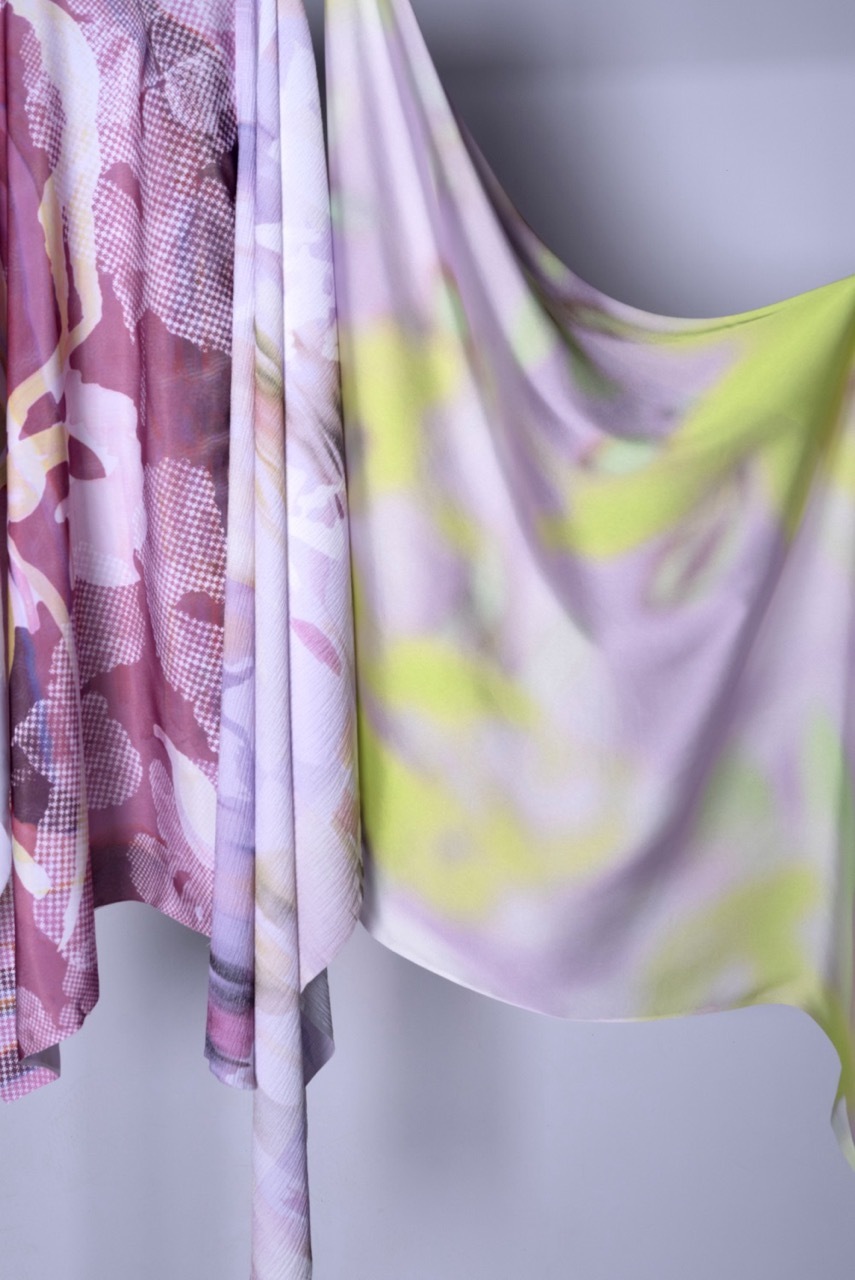 Draped fabrics in pink and white patterns, and pale purple with yellow swirls, hanging against grey background.