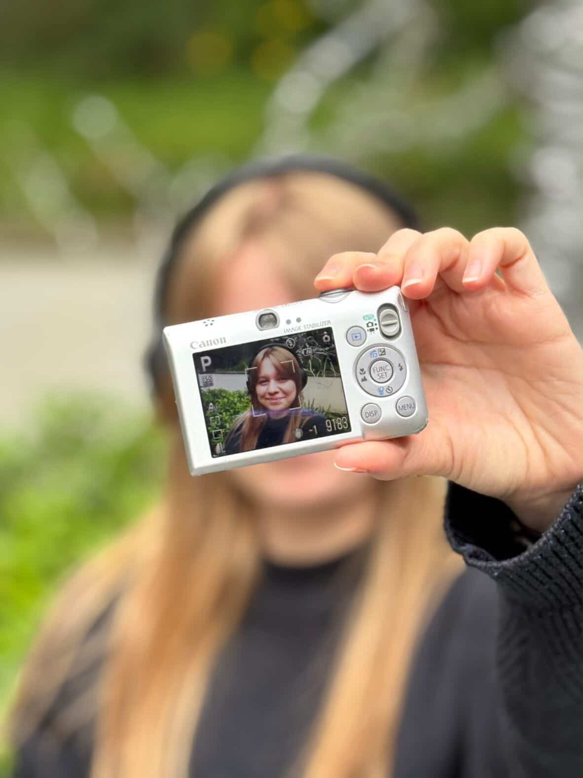E Clements holding a Canon digital camera with a display screen facing forward, showing a photo of a girl with brown hair. The background features blurred greenery and light out of focus.