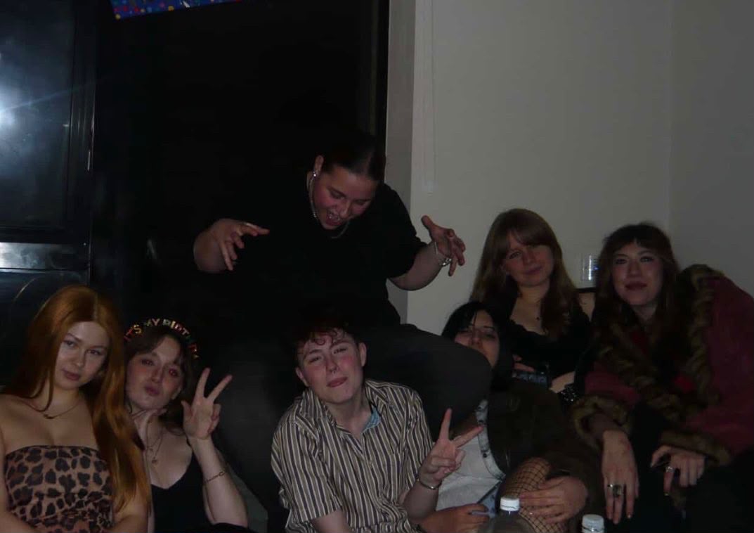 A group of seven individuals seated closely together on the floor of a room. The setting appears informal, with a mix of casual clothing styles. The background includes a large window and soft lighting.