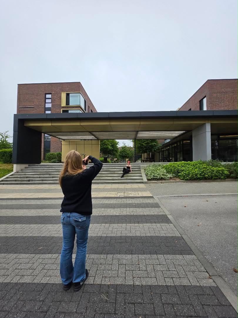 A BA (Hons) Content Creation with Marketing students stands on stone steps on AUB Campus, taking a photograph. In the background, a person sits on the steps of Campus Halls with greenery surrounding.