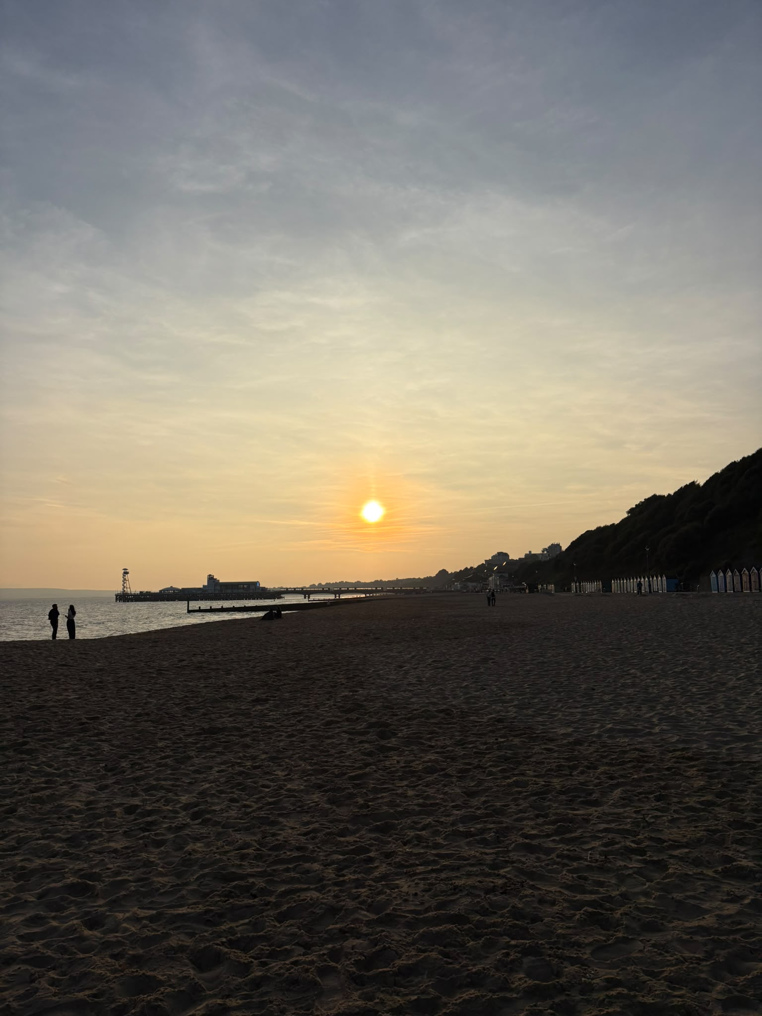 A sandy beach at sunset with the sun just above the horizon. Silhouettes of two people stand on the left, while a pier and coastal trees are visible in the background, creating a serene coastal landscape.