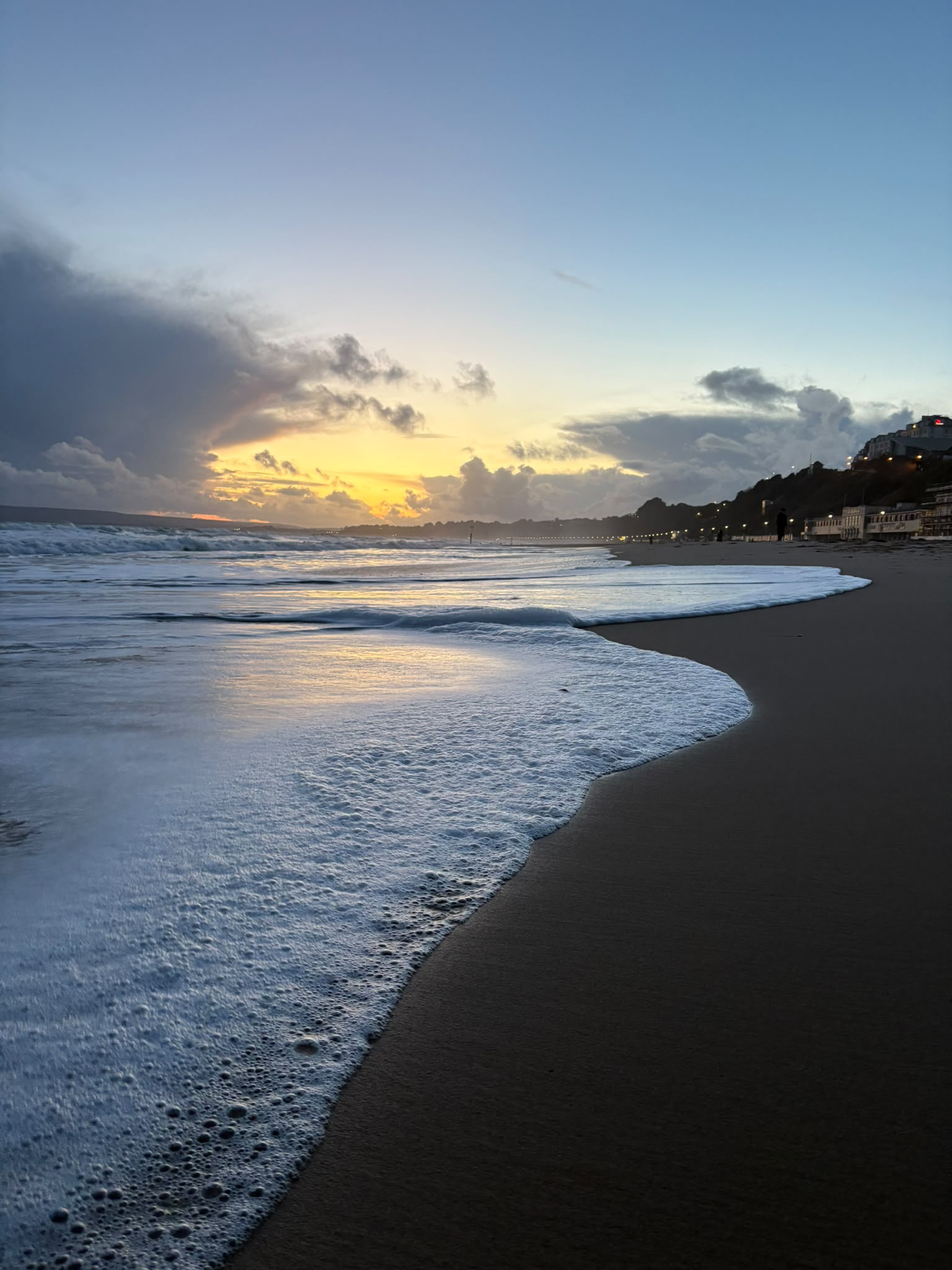 A beach scene at sunset with gentle waves lapping at the shore. The sky features shades of orange and blue while clouds are present in the background. The coastline is visible with buildings on the right and sandy beach in the foreground.