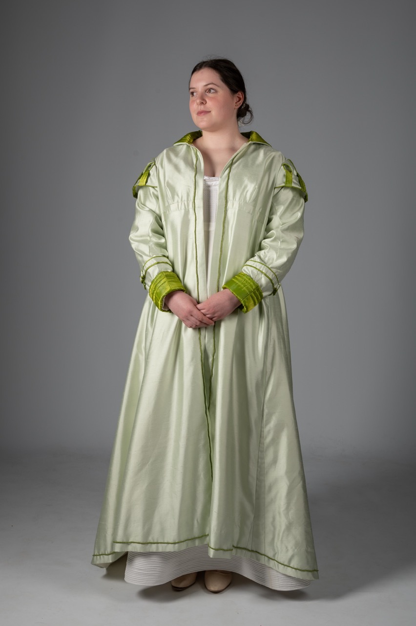 Woman in long pale green hooded robe with darker green trim at cuffs and shoulders, standing against grey background.