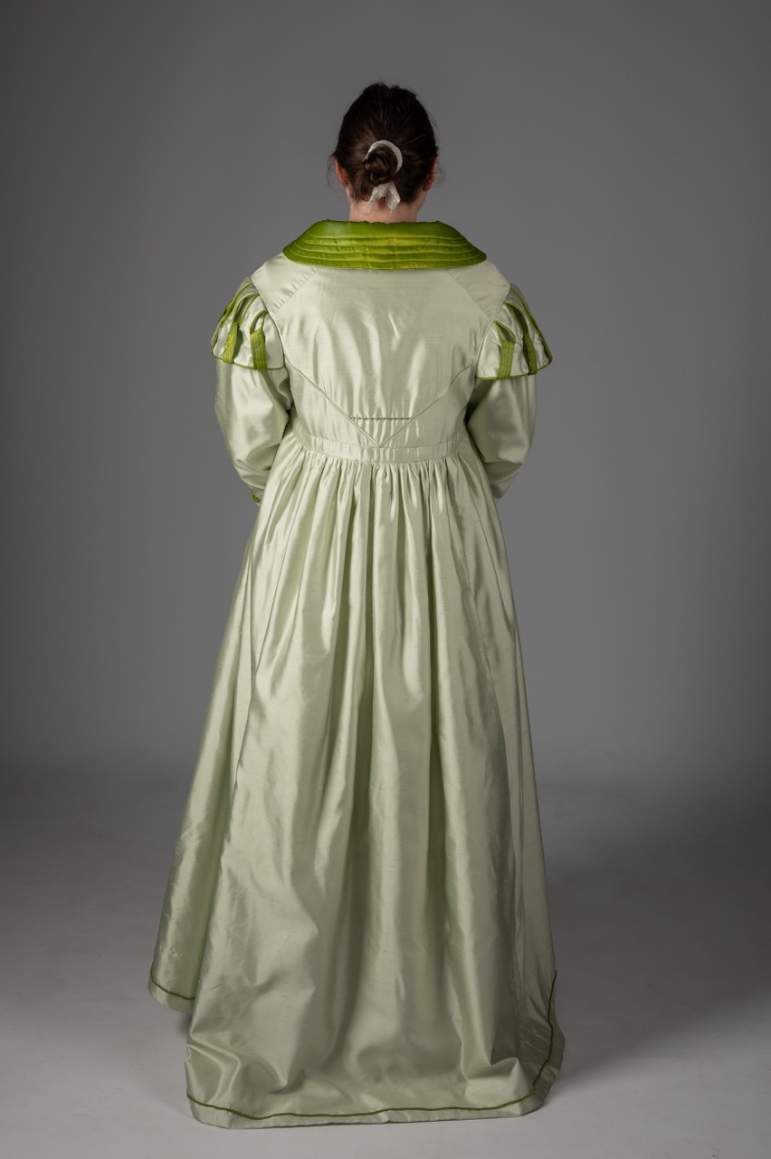 Woman in pale green silk gown with darker green trim, viewed from behind against grey background, hair in low chignon.