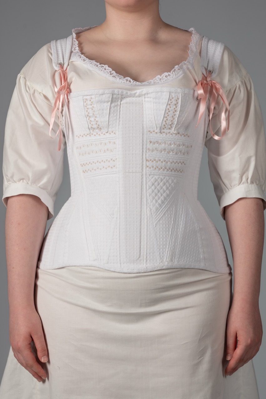 Woman wearing white Victorian-style corset with lace trim and pink ribbon bows over cream blouse and skirt against grey background.