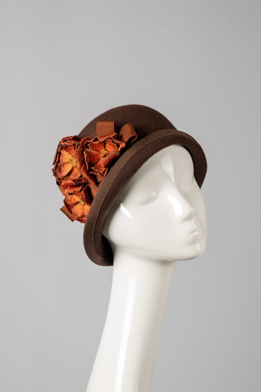 Brown felt hat with orange and red fabric flowers on white mannequin head against grey background.