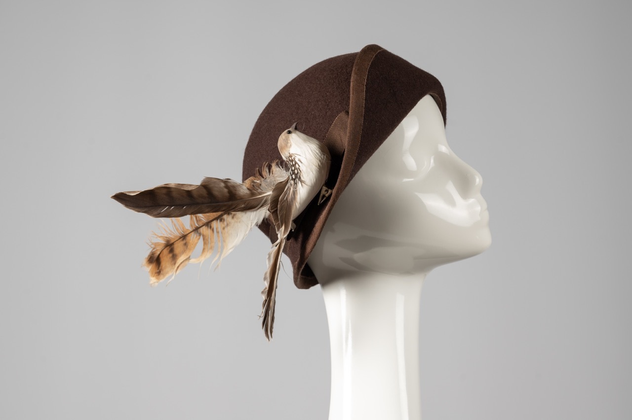 Brown fabric hat with feathers attached, displayed on white mannequin head against grey background.