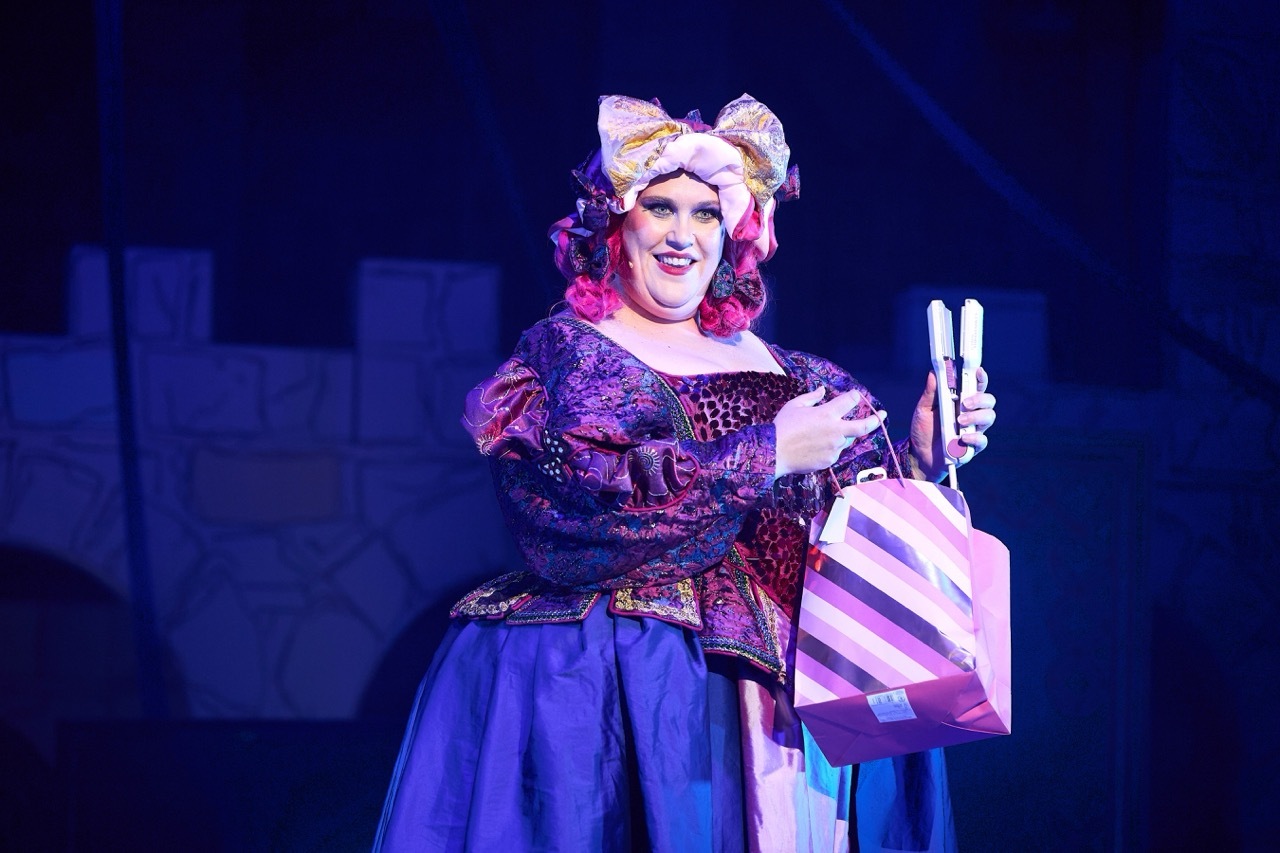 Performer in period costume with white bonnet, burgundy bodice, and blue skirt holds striped shopping bag on blue-lit stage.