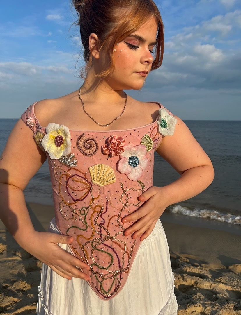 Woman wearing pink embroidered top with floral and shell motifs, white skirt, standing on beach with blue sky and sea behind.