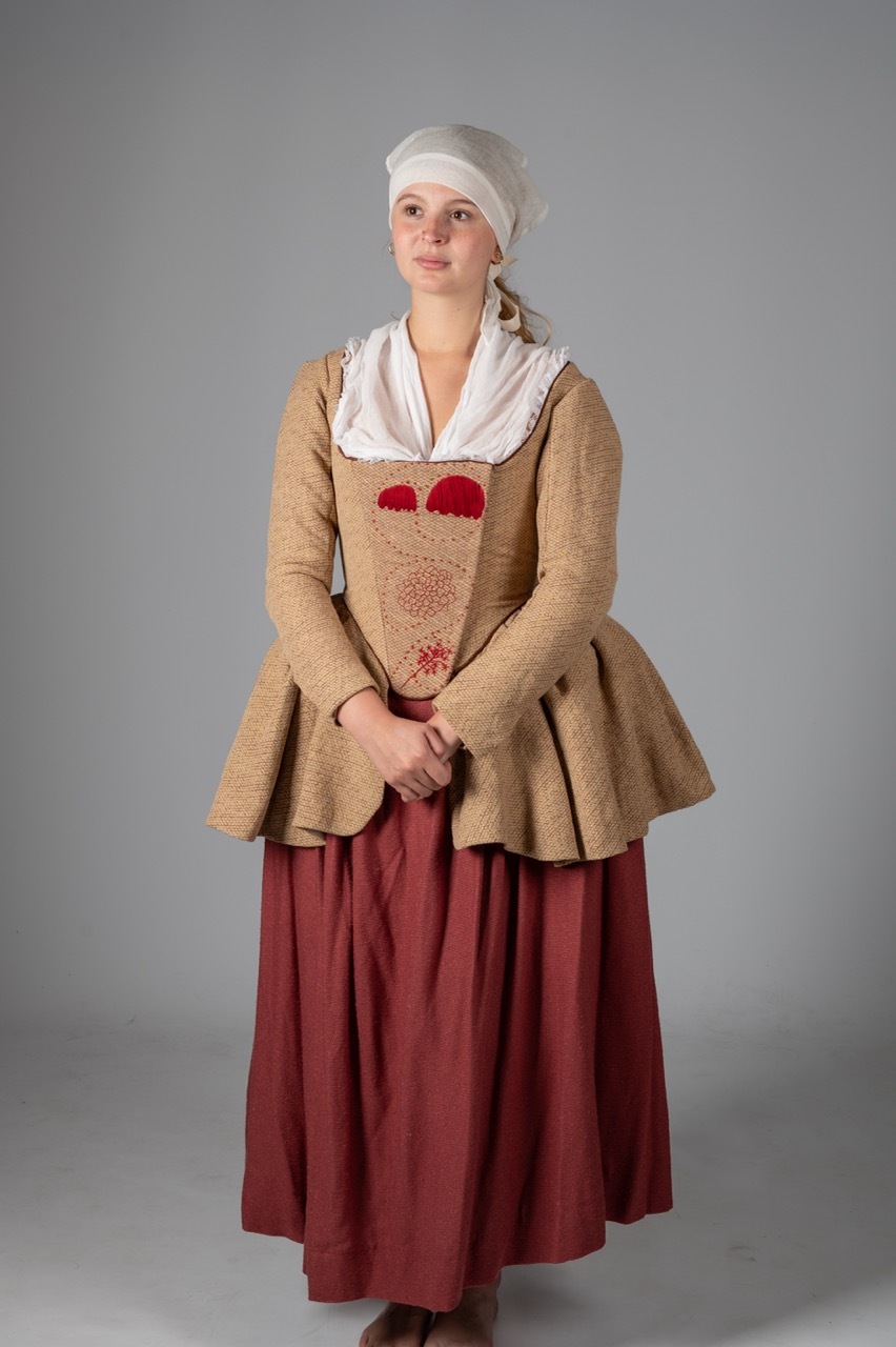 Woman in historical costume with white cap, beige bodice with red embroidery, white collar, and burgundy skirt against grey background.