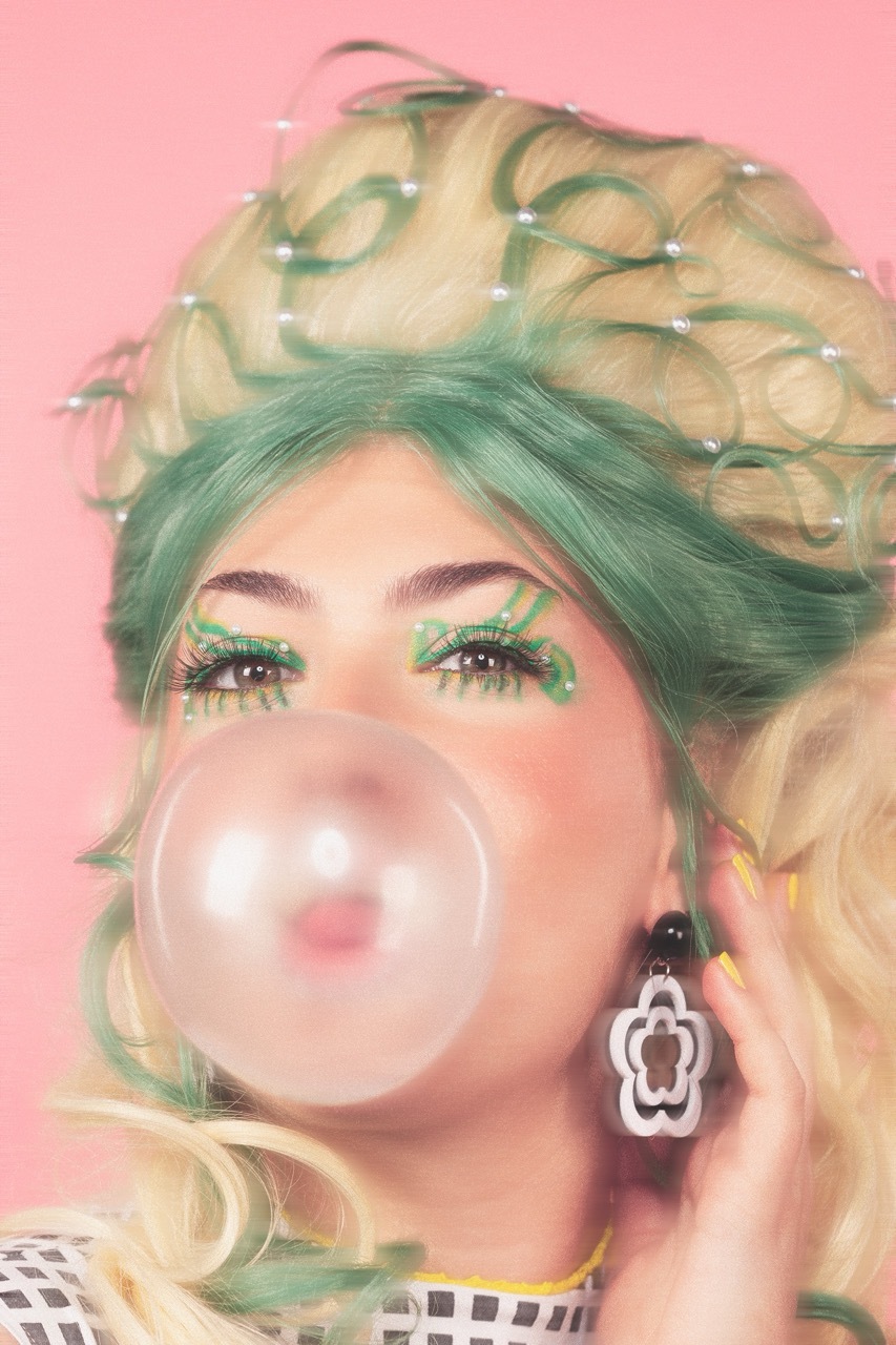 Woman with green-tinted blonde hair and matching eye makeup blowing a bubble against pink background, wearing white earrings.