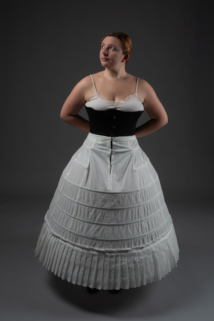 A woman stands in a fitted black corset paired with a long white dress featuring pleated layers. The background is grey, highlighting the dress and its structure.