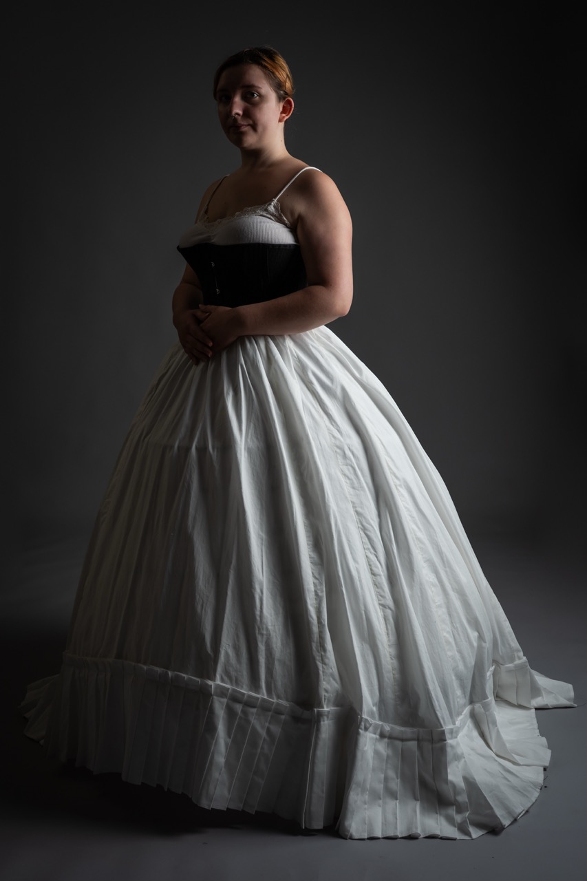 A woman dressed in a floor-length white gown with a fitted black bodice stands against a dark background. The gown features a full skirt with pleats and a ruffled hem, showcasing the elegant silhouette of the outfit.