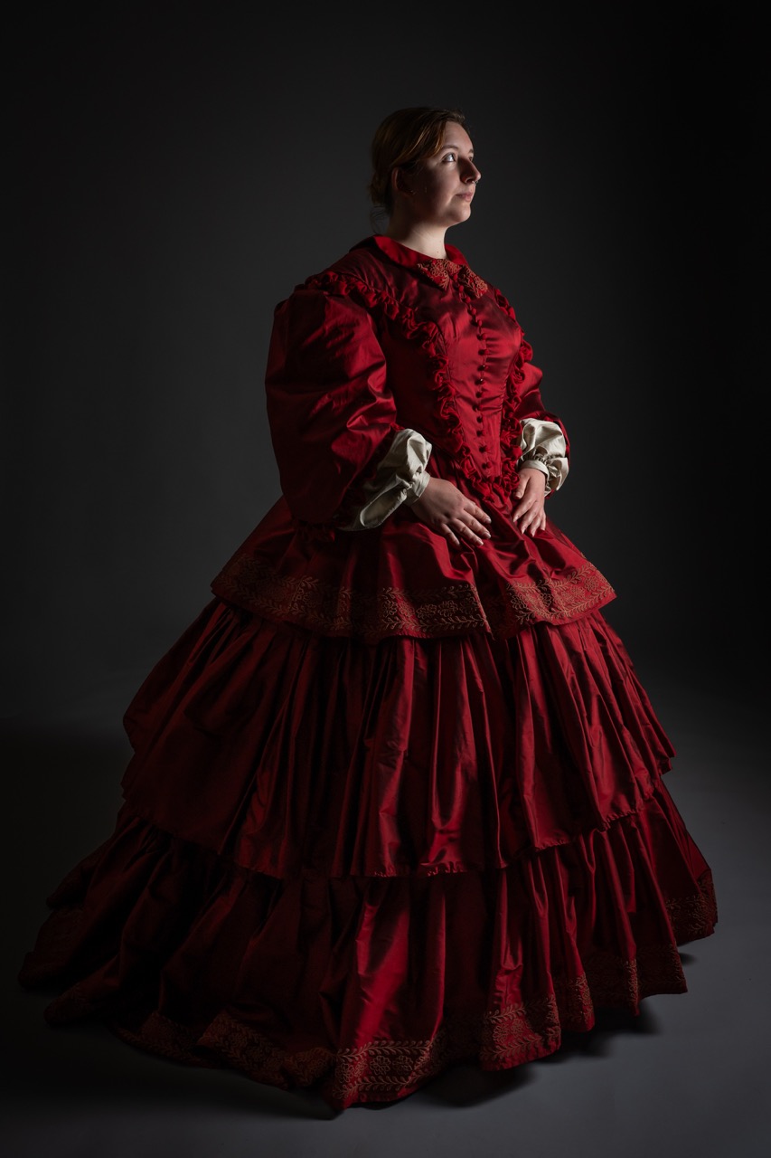 A woman wearing a voluminous red Victorian gown with layered ruffles stands in a dimly lit space. The dress has a high neckline and ornate detailing, contrasting with the dark background.