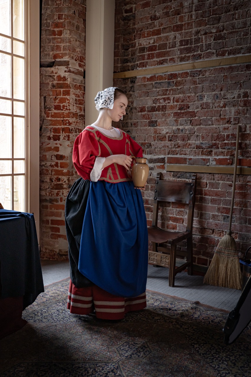 A woman in a historical costume stands in a room with exposed brick walls. She wears a red blouse with white accents and a long blue skirt, holding a clay jug. A wooden chair, broom, and a patterned rug are visible in the background.