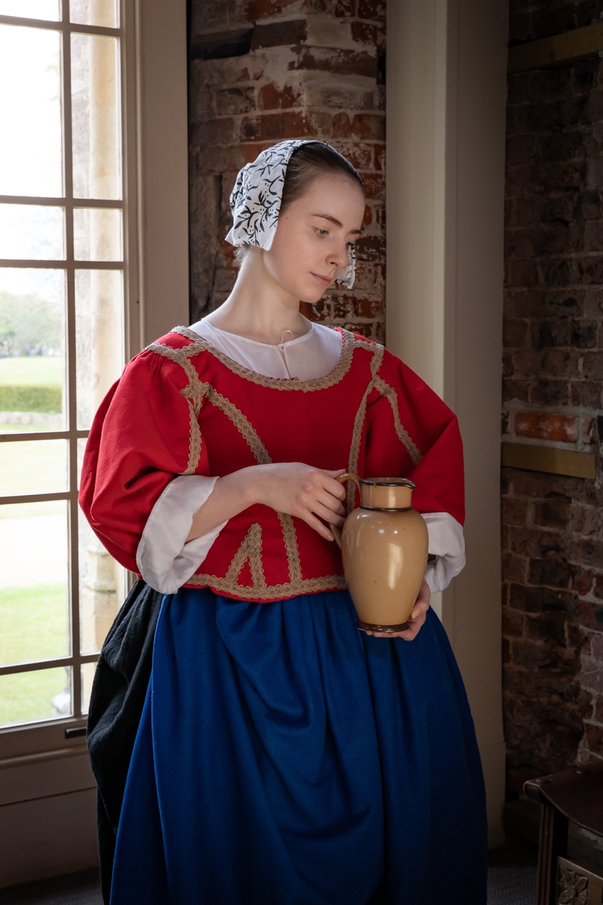 A young woman in historical clothing stands beside a large window. She wears a red bodice with gold trim over a blue skirt, and holds a clay jug in her hands. A stone wall is visible behind her.