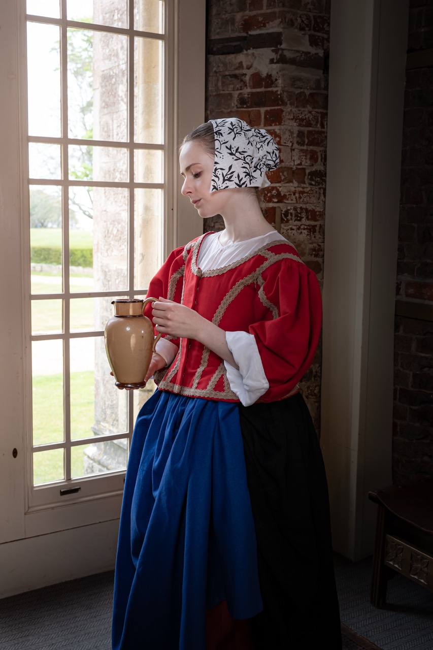 A woman in historical attire holds a clay jug while standing beside a large window. She wears a red top adorned with gold trim, a white blouse, and a blue skirt, and her head is covered with a patterned white bonnet. The background features brick walls and greenery visible outside.