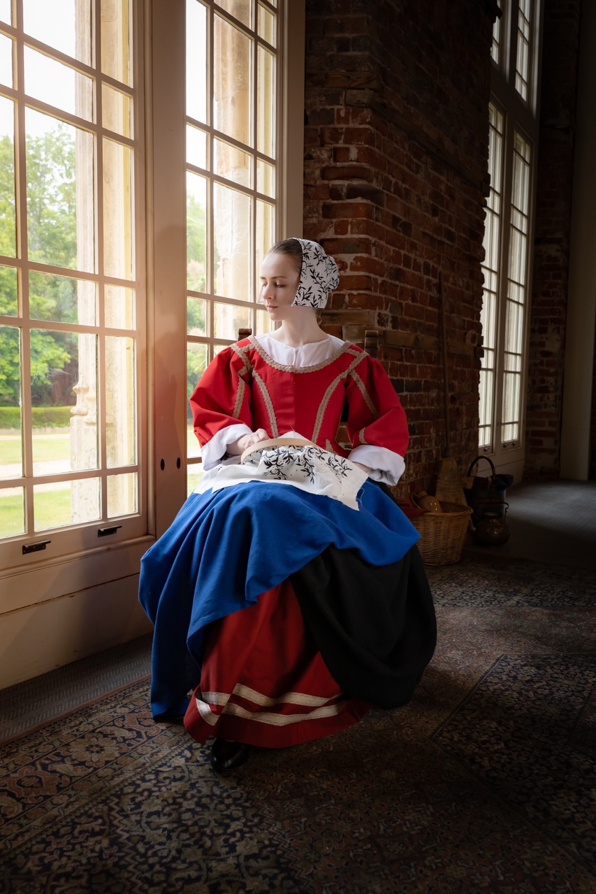 A woman dressed in a historical costume with a red and blue gown is seated by large windows. The setting features exposed brick walls and a patterned rug, while natural light illuminates the space and reveals greenery outside.