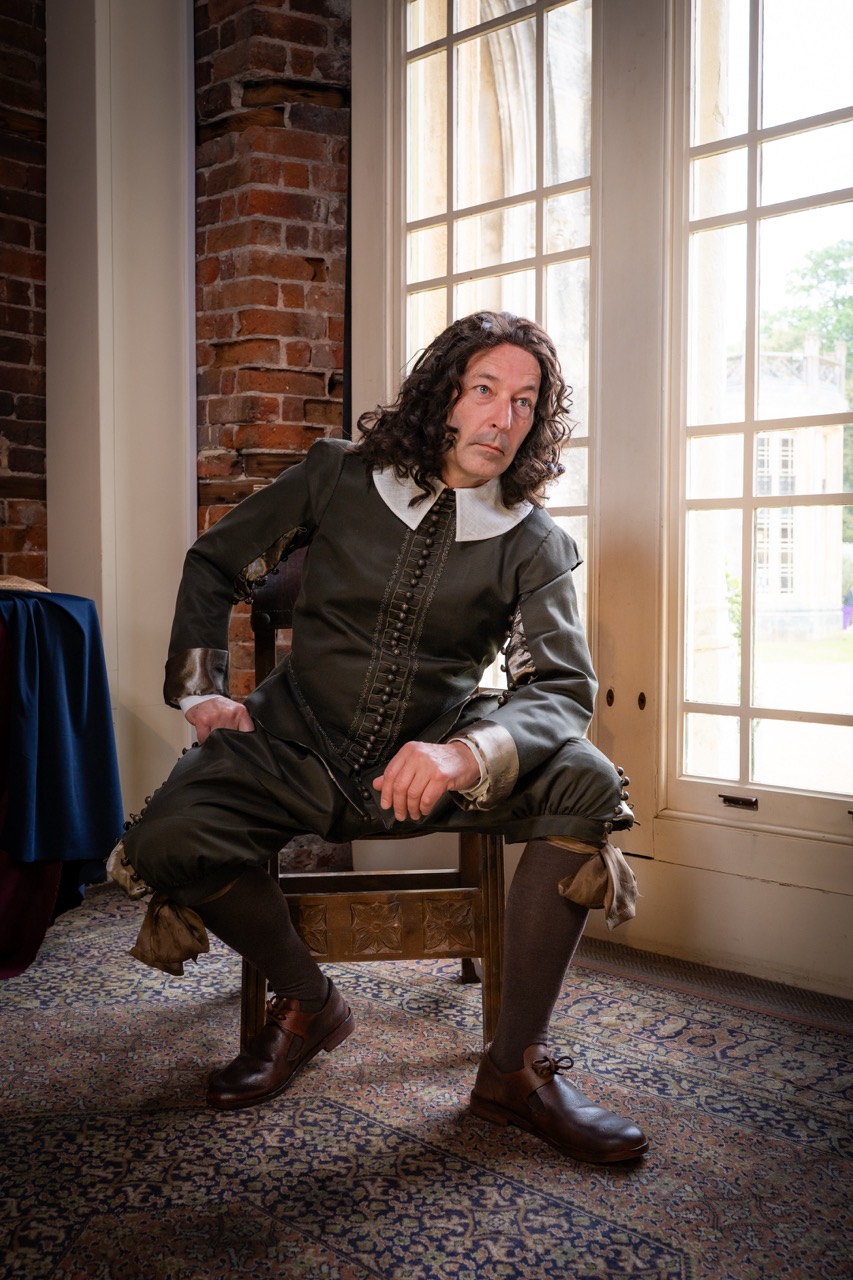 A man dressed in 17th-century attire, featuring a dark coat with a white collar, sits on a wooden chair near large windows. The room has brick walls and a patterned carpet, creating a historical atmosphere.