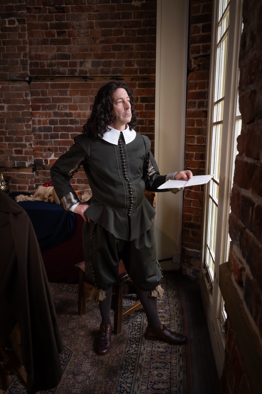A man dressed in a historical dark green outfit with a white collar stands by a window, holding a piece of paper. The background features a brick wall and a table with various items, illuminated by natural light.