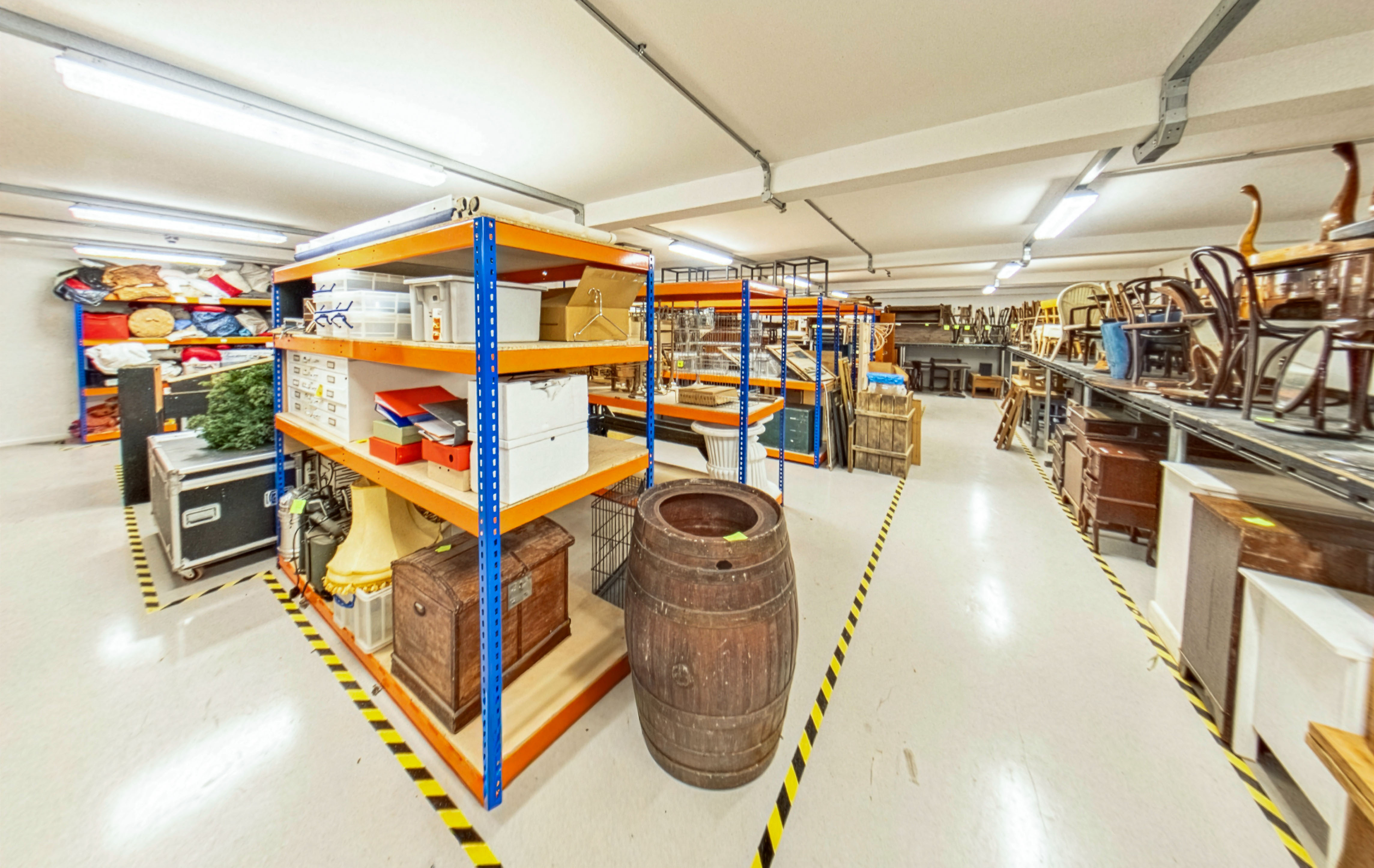 Large warehouse storage area with blue and orange metal shelving units containing boxes and items, yellow striped floor markings.