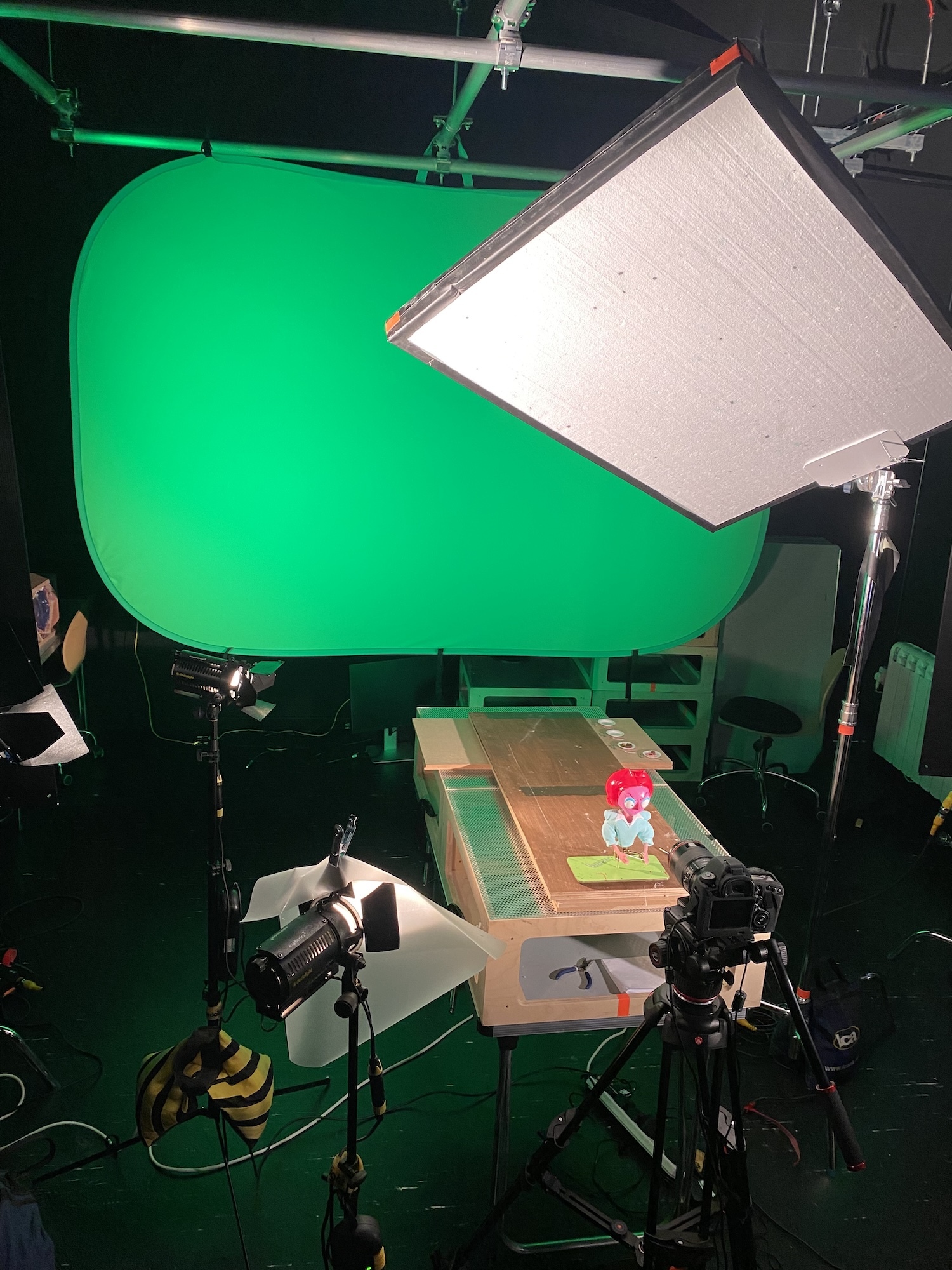 Stop motion animation studio with green screen backdrop, white lighting panel, camera on tripod, and miniature set with cardboard buildings below.