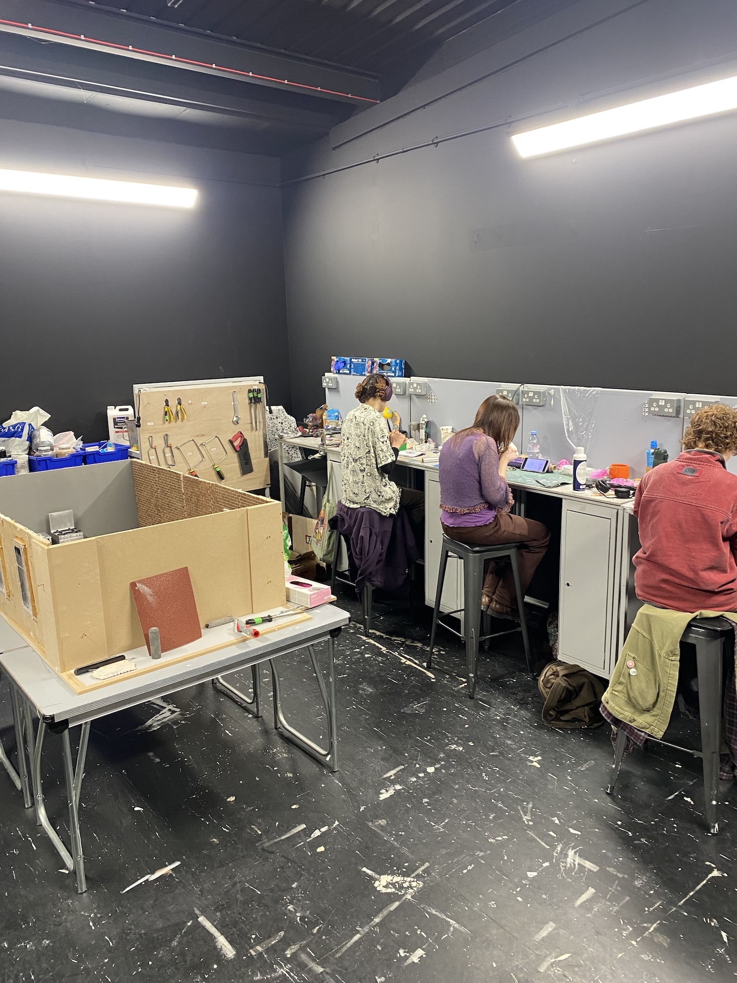 Three people working at tables in art studio with grey walls, exposed ceiling, fluorescent lighting, and cardboard materials scattered about.