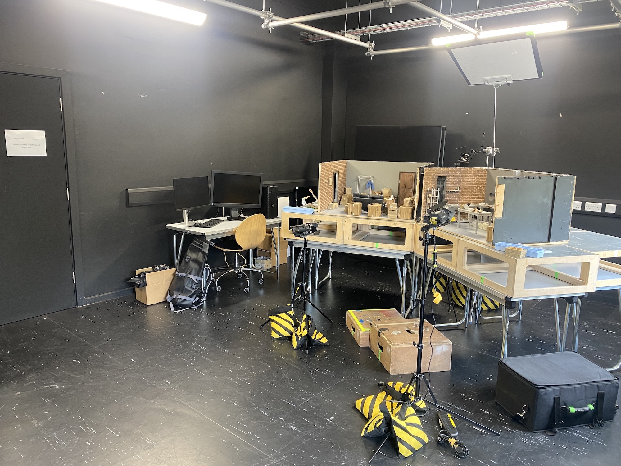 Dark grey studio space with wooden work tables, cardboard boxes, black chairs, and yellow/black striped materials scattered on floor.
