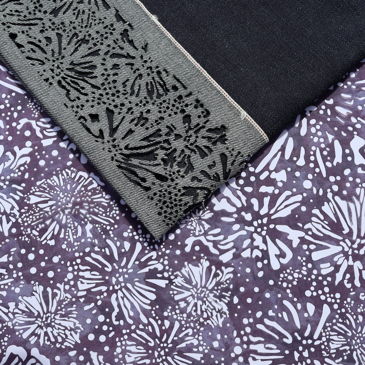 A close-up of two fabric swatches: one features a dark black lace pattern along the edge, while the other displays a floral print in shades of purple and white. The fabrics are layered, showcasing their contrasting designs and textures.