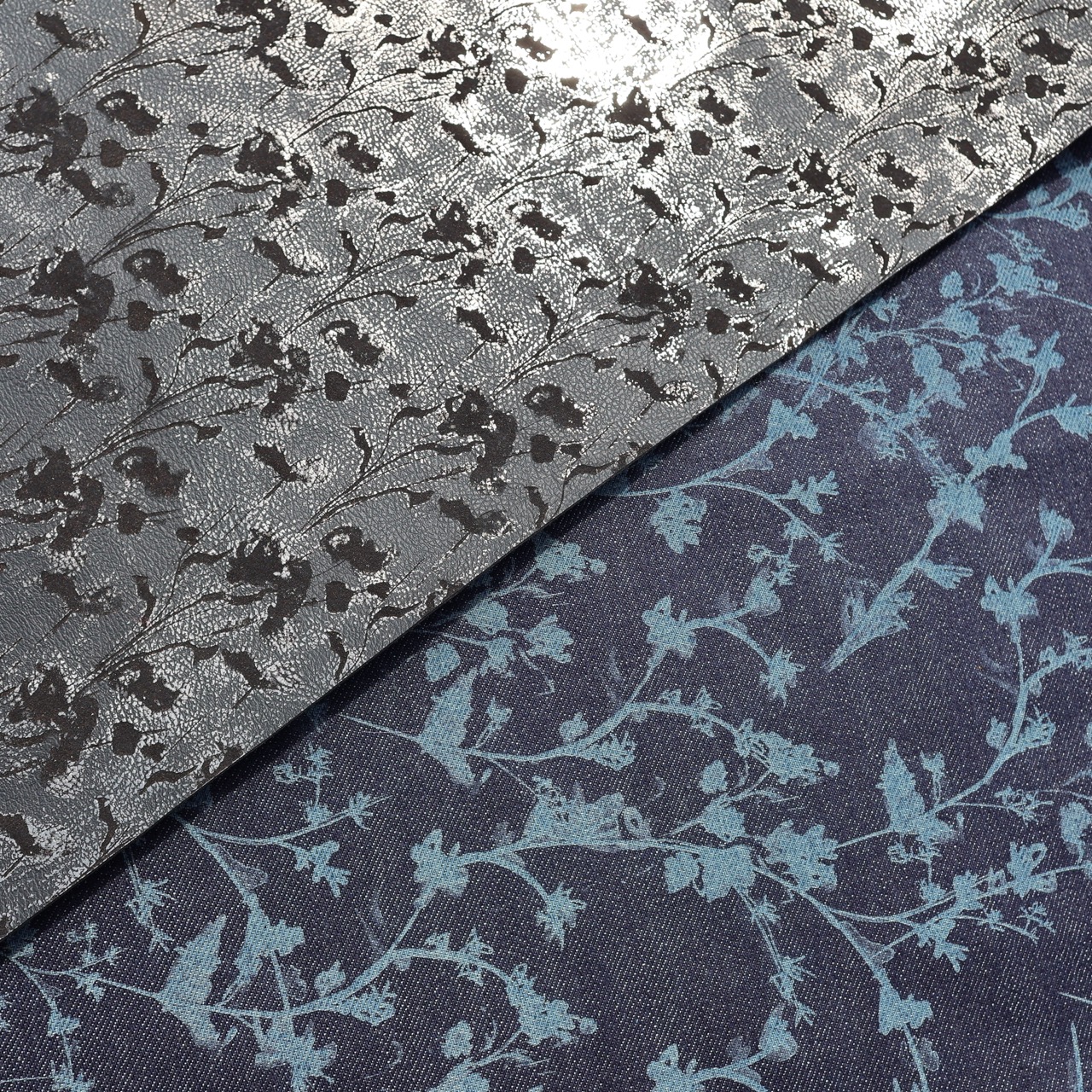 A close-up of two pieces of fabric laid side by side. The top fabric is grey with a shiny finish featuring a delicate floral pattern, while the bottom fabric is a dark blue with a softer, muted floral design in light blue.