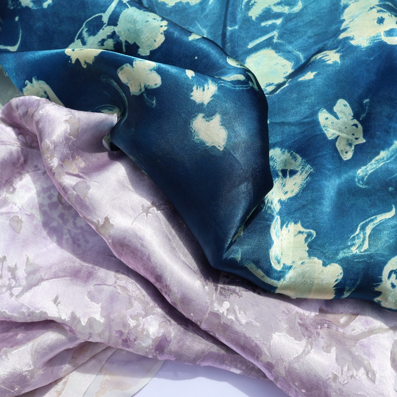 Fabric pieces in varying colours and patterns, including a dark blue silk with light floral designs and a light purple fabric with a subtle floral pattern. The textures appear soft and slightly transparent.