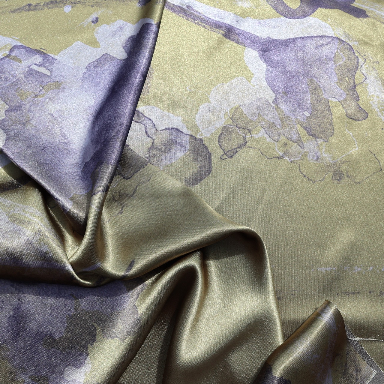 A folded piece of fabric displaying an abstract floral pattern in shades of purple and cream against a muted olive background. The smooth texture of the satin fabric catches the light, emphasizing the flowing folds.