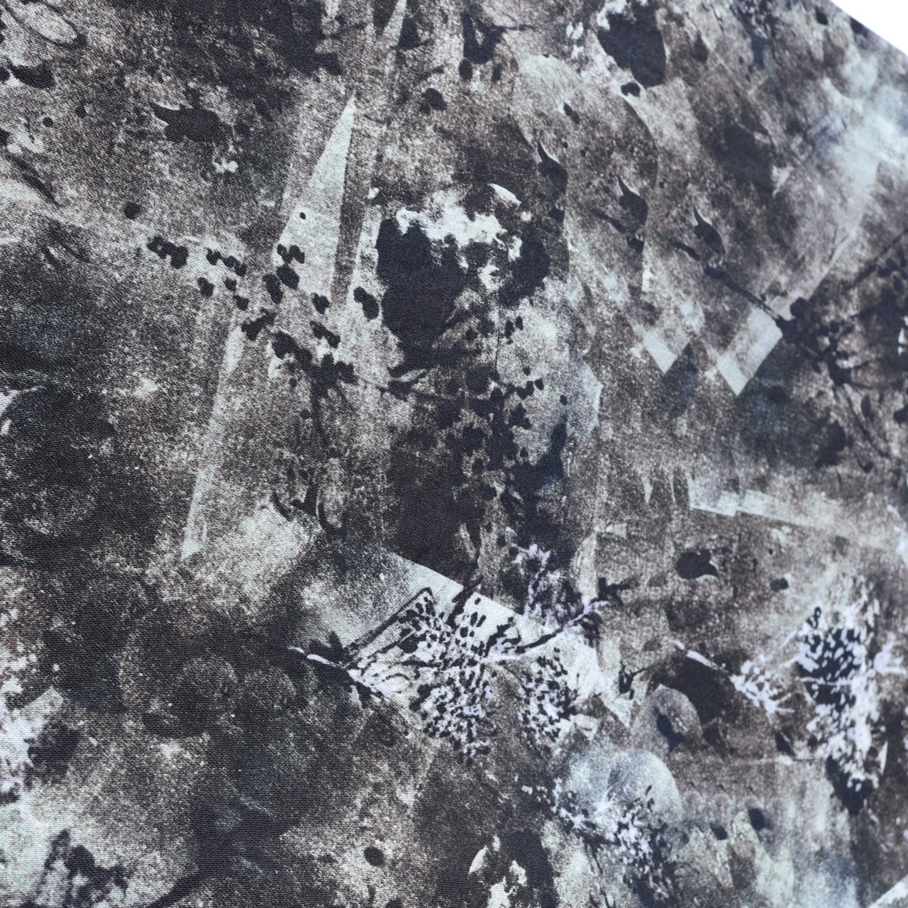 A textured fabric surface in shades of black, grey, and white featuring a chaotic pattern of splotches and abstract shapes. The design includes both dark spots and lighter areas, creating a visually striking effect.