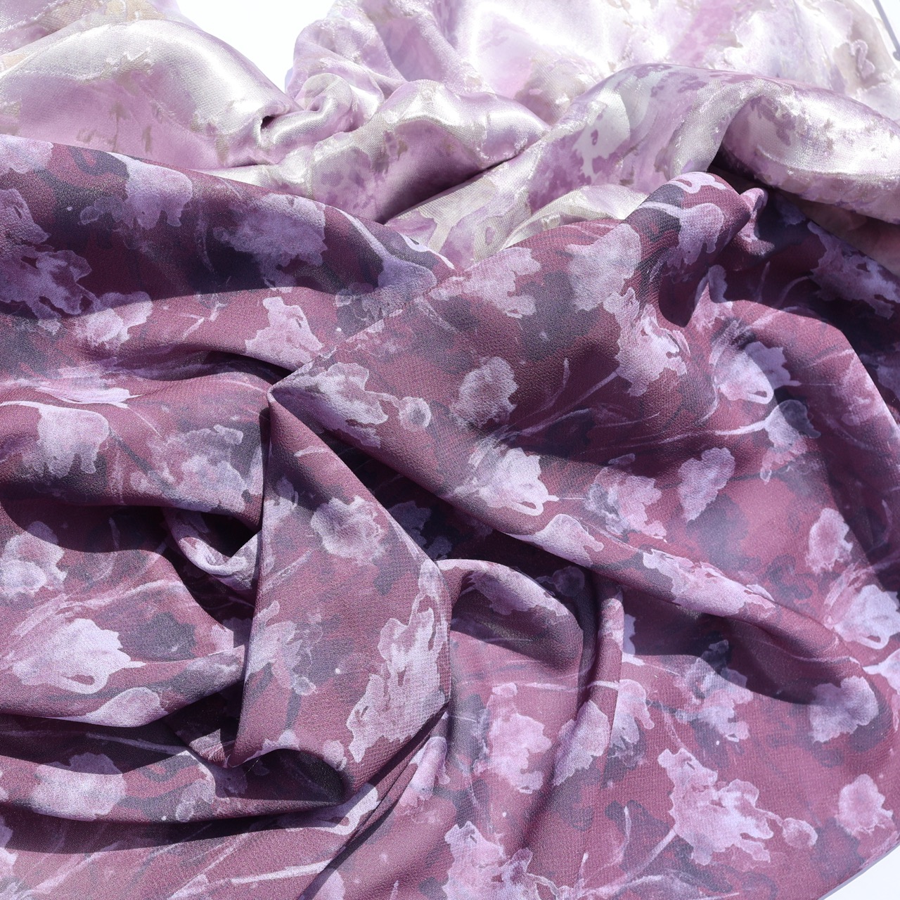 A folded piece of fabric in shades of purple, featuring a floral pattern with soft, blurred details. The texture appears lightweight and slightly sheer, showcasing a flowing drape with subtle variations in light and colour.