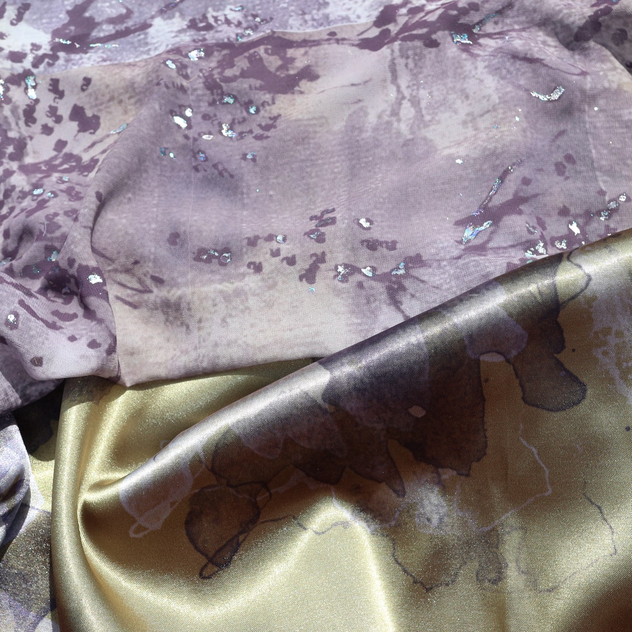 A textured fabric featuring a blend of soft purple tones with sequins on top, layered over a shiny, golden silk fabric displaying a subtle pattern of darker shades. The fabrics are arranged in a draped manner.